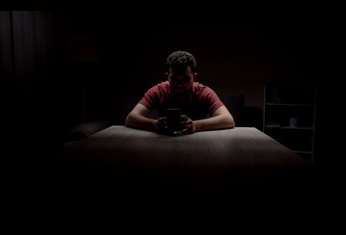 Person sitting alone in a dark room illuminated by a phone screen, representing pornography addiction, isolation, and mental health struggles
